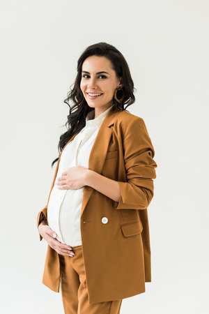 Blissful pregnant woman in brown suit touching tummy and looking at camera isolated on whiteの写真素材