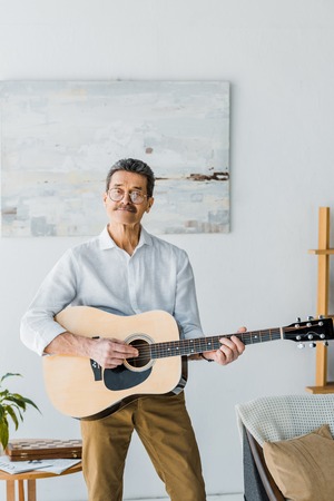 happy senior man in glasses playing acoustic guitar at home