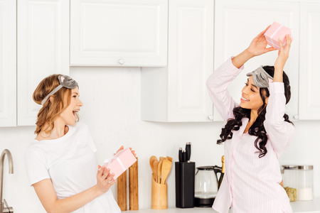 beautiful happy multicultural girls in sleeping masks and pajamas with gift boxes in kitchenの写真素材