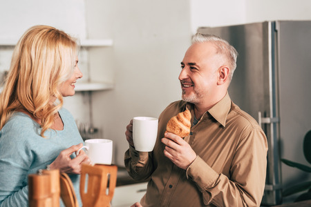 cheerful man holding tasty croissant and cup of drink while looking at wifeの写真素材