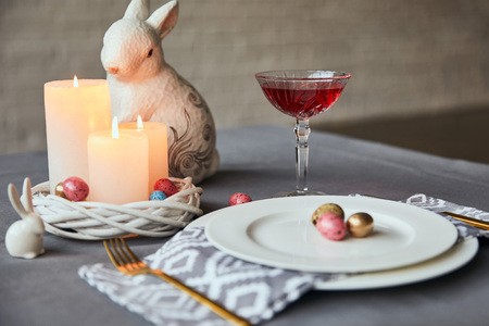selective focus of plates with napkin, eggs and burning candles in basket, wine in crystal glass and decorative rabbit on table at homeの写真素材