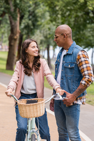 handsome african american man standing near cheerful girlfriend riding bicycleの写真素材