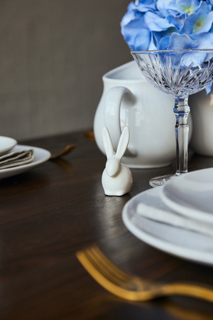 selective focus of decorative rabbit near plates and crystal glass on wooden table at home