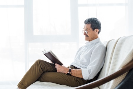 thoughtful senior man reading book while sitting on sofaの写真素材
