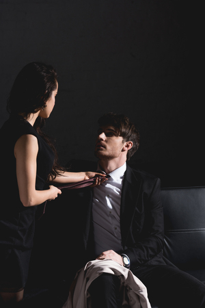 brunette woman in black dress taking off tie with handsome sitting on couch on dark backgroundの写真素材