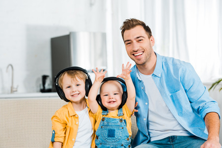 smiling father looking at camera with adorable toddler and preschooler sons in headphones at home