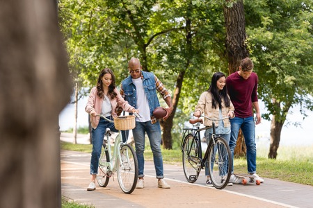 african american man walking near pretty girls riding bicycles and cheerful friend longboarding in...