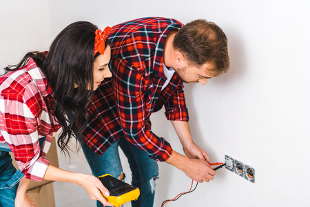cheerful woman holding digital multimeter near boyfriend looking at power socketの写真素材