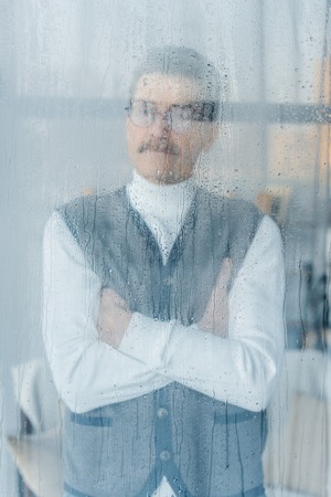 pensive retired man standing with crossed arms near windowの写真素材