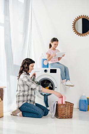 daughter in t-shirt sitting on washer with digital tablet wile mother in grey shirt taking clothes from basket in laundry roomの写真素材