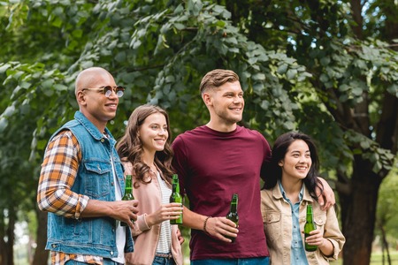happy multiethnic group of friends holding bottles with beer in park