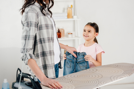 cropped view of mother and daughter holding jeans near ironing board at homeの写真素材
