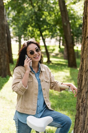 happy young woman in sunglasses talking on smartphone near bike in park