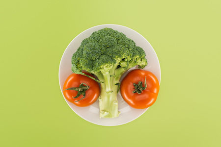 top view of organic ripe broccoli near red tomatoes on white plate isolated on greenの写真素材