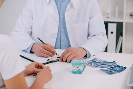 cropped view of orthodontist writing diagnosis during consultation with woman in clinic