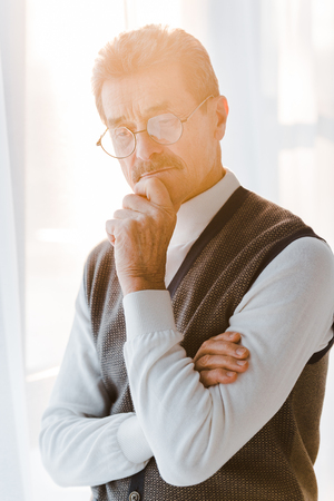 pensive senior man with grey hair standing with crossed arms at homeの写真素材