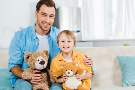 father and preschooler son sitting on couch and holding teddy bears at homeの写真素材