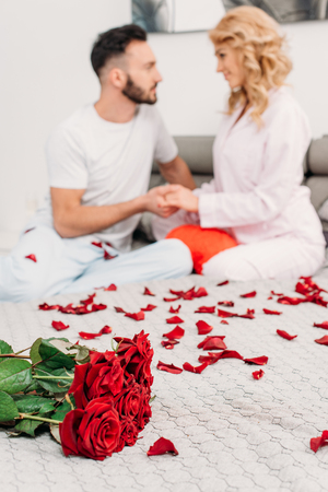 Couple sitting on bed with red roses and holding handsの写真素材