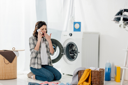 woman sitting near washer, talking on smartphone and yawning in laundry roomの写真素材