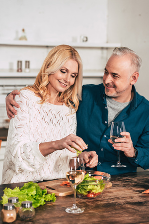 handsome man with glass of wine hugging attractive blonde wife holding green lettuce leaves