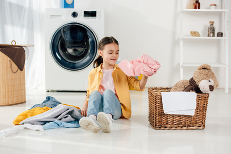 child in yellow shirt and jeans sitting and putting clothes to basket in laundry roomの写真素材