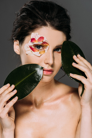 beautiful young woman with floral petals around eye holding leaves isolated on greyの写真素材