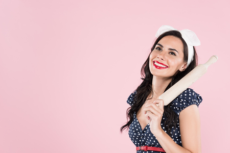 Laughing brunette woman in dotted dress holding wooden rolling pin isolated on pinkの写真素材