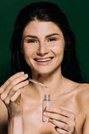 cheerful naked girl holding bottle with moisturizing serum isolated on greenの写真素材