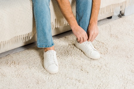 cropped view of man in jeans sitting and tying shoelaces on sneakers at home