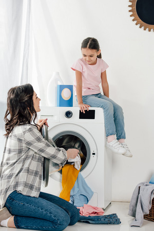 daughter in pink t-shirt sitting on washer wile mother in grey shirt putting clothes in laundry roomの写真素材