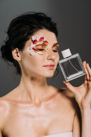 tender girl with closed eyes and petals on face holding perfume bottle isolated on greyの写真素材