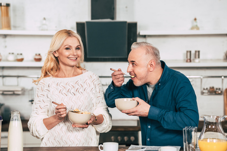 attractive wife holding bowl with cornflakes near cheerful husband eating in kitchenの写真素材