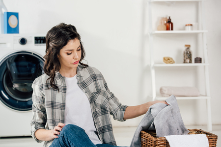 selective focus of attractive woman sitting on floor and putting clothes to basket in laundry roomの写真素材