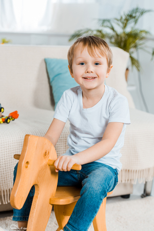 adorable smiling preschooler boy riding rocking horse at home and looking at cameraの写真素材