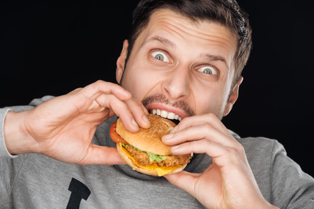 emotional man eating tasty chicken burger while looking at camera isolated on blackの写真素材