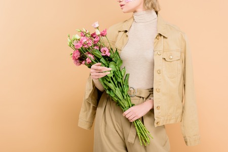cropped view stylish woman with flowers on face holding bouquet isolated on beige