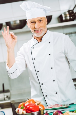 handsome smiling male chef in uniform looking at camera and showing ok sign in restaurant kitchenの写真素材