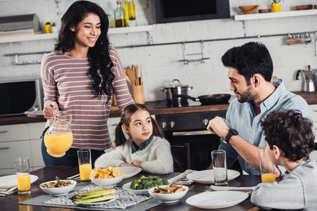 cheerful latin mother holding jar with orange juice near daughter, husband and son during lunchの写真素材