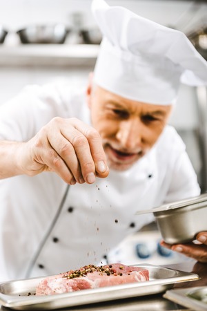 male chef in uniform seasoning meat in restaurant kitchen