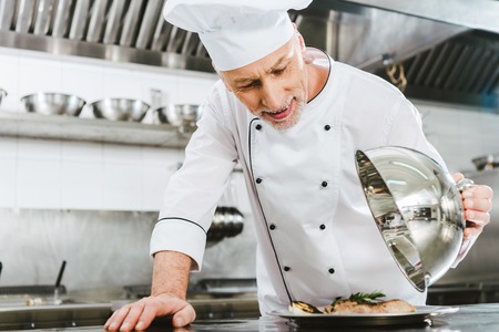 selective focus of handsome male chef in uniform holding dome from serving tray with meat dish in...