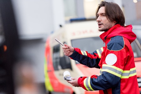 Brunette paramedic in red uniform holding pen and clipboard on streetの写真素材