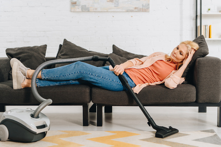Tired senior woman with vacuum claner lying on sofaの写真素材