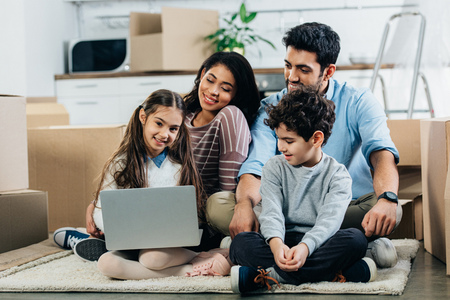 cheerful hispanic family looking at laptop while sitting on carpet in new homeの写真素材