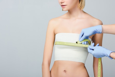cropped view of doctor holding measuring tape on female breast with bandage after plastic surgery, isolated on greyの写真素材