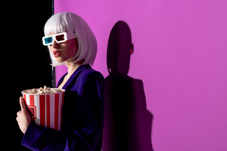Interested girl in 3d glasses holding popcorn on purple background