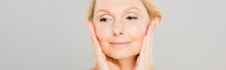 panoramic shot of attractive and smiling mature woman touching her face and looking away on grey...