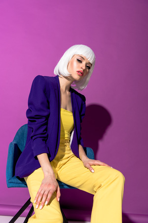 Fashionable woman in white wig sitting in armchair on purple backgroundの写真素材