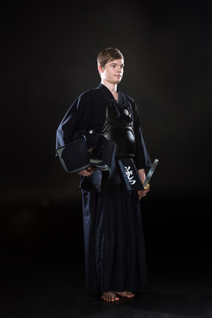 Full length view of young man holding kendo helmet and bamboo sword on blackの写真素材