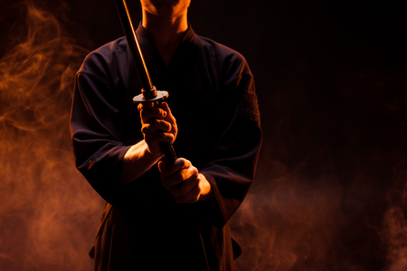 Cropped view of young man in kimono holding kendo sword in smokeの写真素材