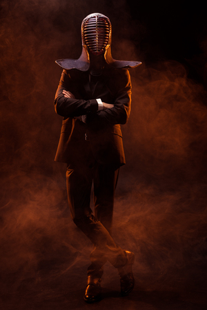 Full length view of kendo fighter in formal wear and helmet standing with crossed arms on darkの写真素材